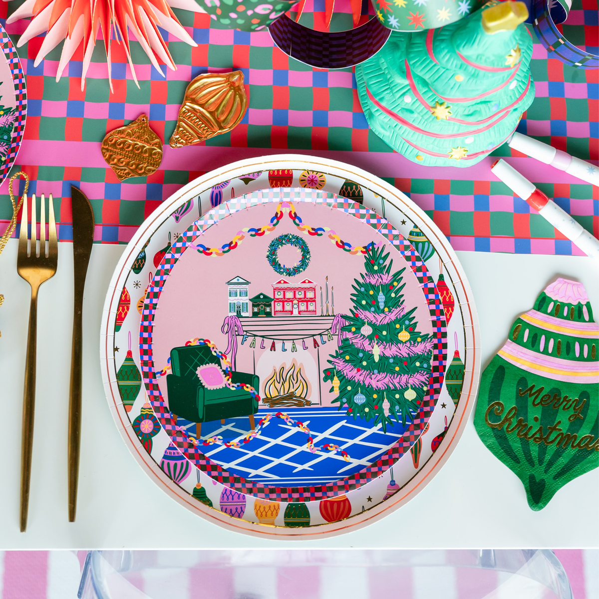 Overhead holiday place setting showing the ornament napkin with ornament-patterned plates, a Christmas tree plate, gold flatware, and a paper fan.