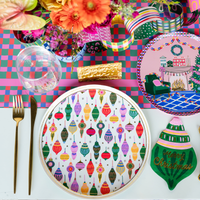 Overhead holiday place setting showing the ornament plate with an ornament-shaped napkin, a green ornament plate, gold flatware, and vibrant Christmas décor on a plaid runner.