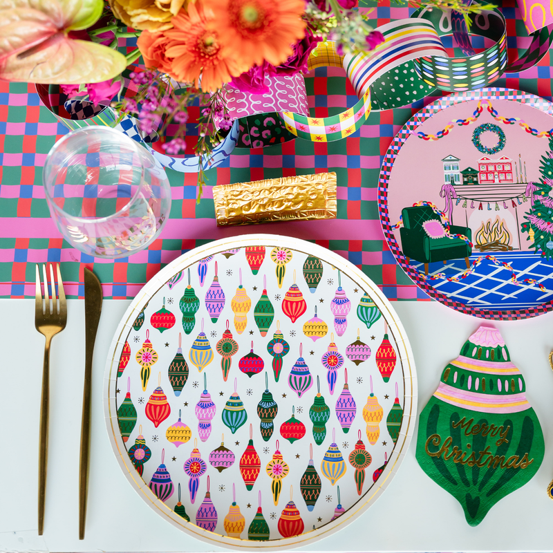 Overhead holiday place setting showing the ornament plate with an ornament-shaped napkin, a green ornament plate, gold flatware, and vibrant Christmas décor on a plaid runner.