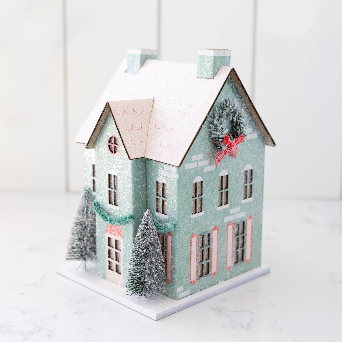 Angled view of a mint putz house showing a frosted wreath with a red bow on the side gable