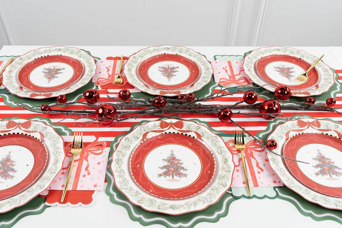 Overhead holiday table with multiple Christmas-tree medallion settings, green scalloped chargers, a striped runner, red ornaments, and gold cutlery.