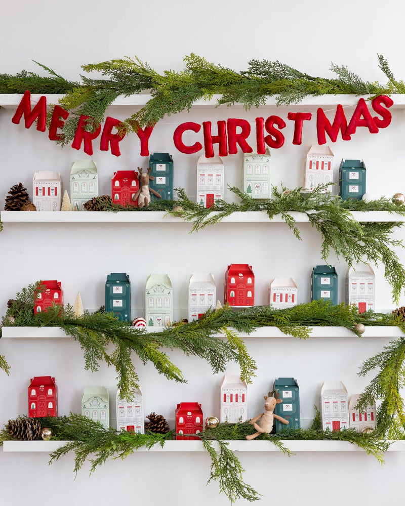 Christmas Village advent boxes styled on a shelf beneath a Merry Christmas banner with garland