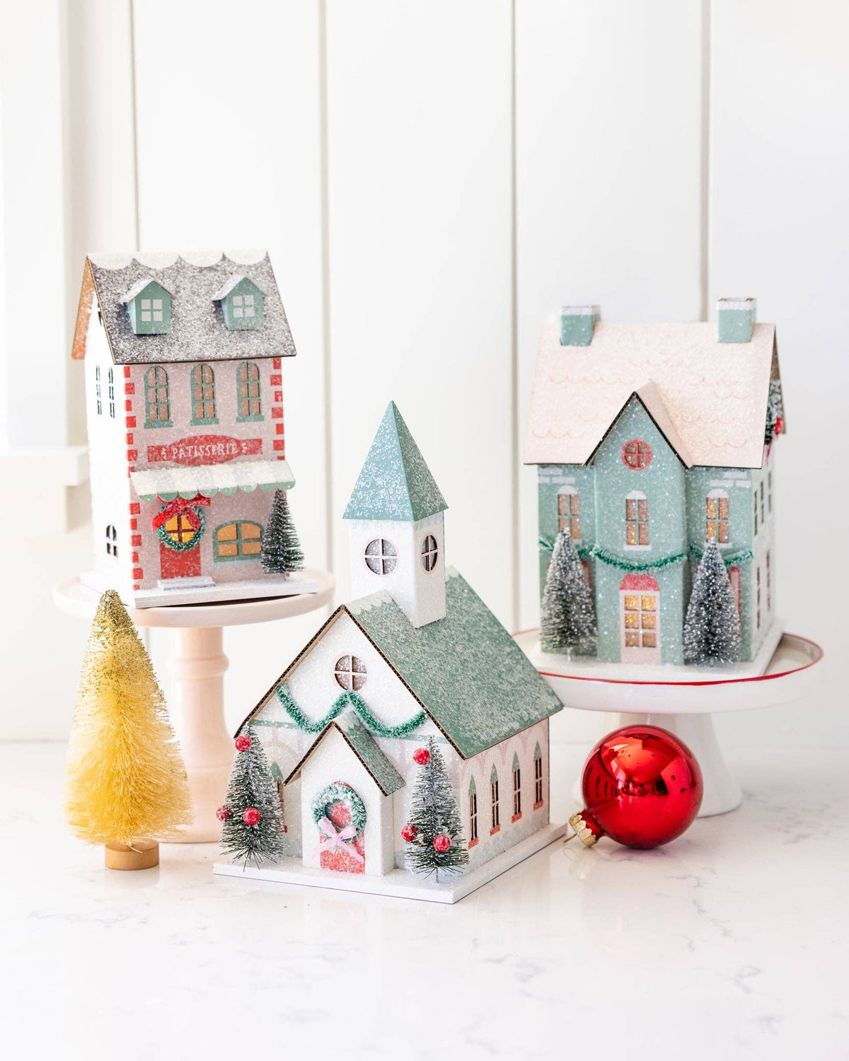 Three glittered Christmas village buildings styled on cake stands: a patisserie, a church, and a mint house