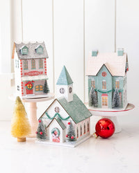 Three glittered Christmas village buildings styled on cake stands: a patisserie, a church, and a mint house