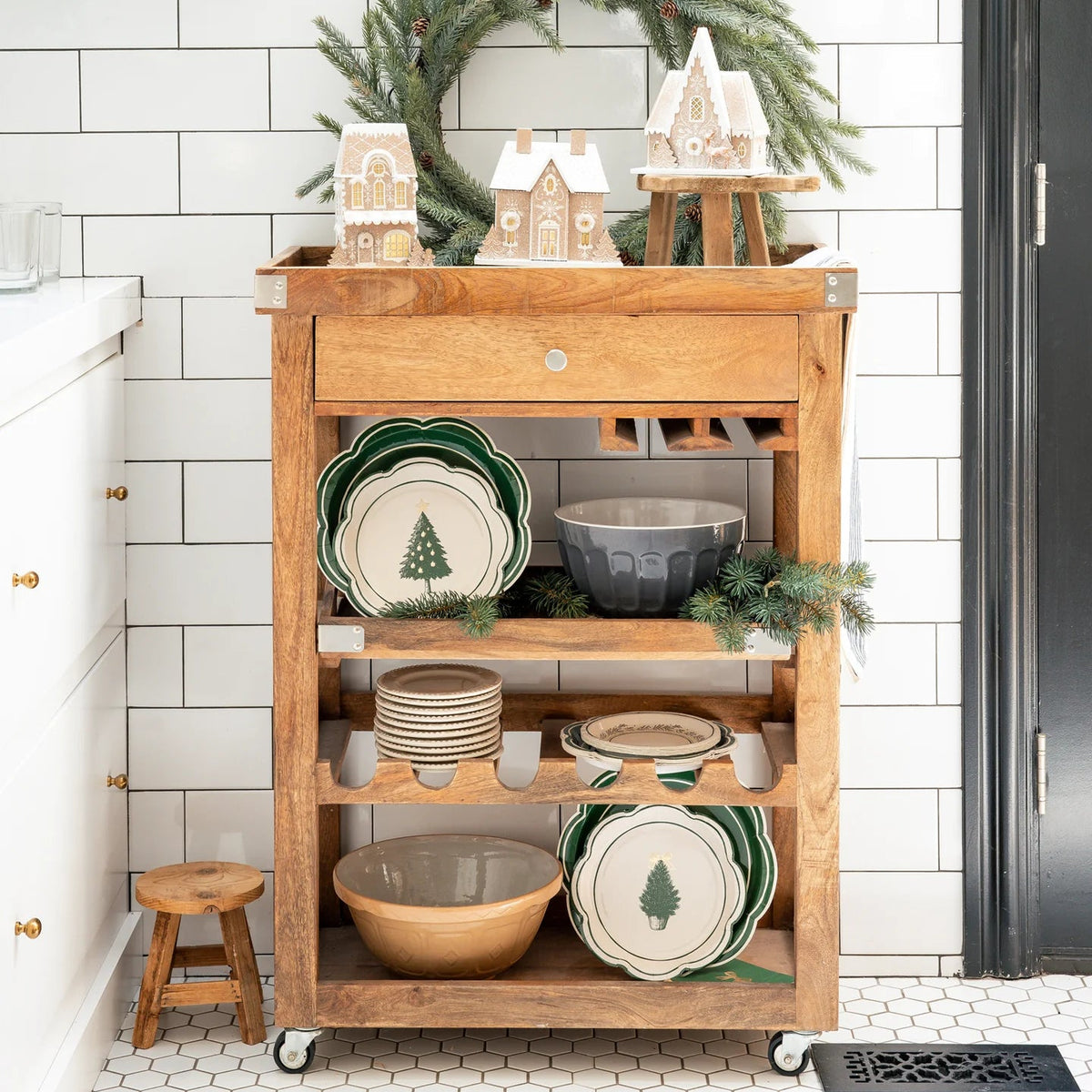 Christmas paper plates stacked on a kitchen bar cart next to gingerbread houses and a wreath, showcasing the disposable holiday tableware.