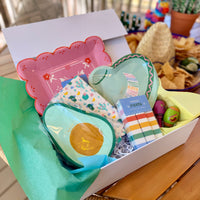 Cinco de Mayo Fiesta Party Kit box open showing all five products in green and blue tissue paper including sombrero plates, pink papel plates, avocado napkins, cacti napkins, and striped fan banner