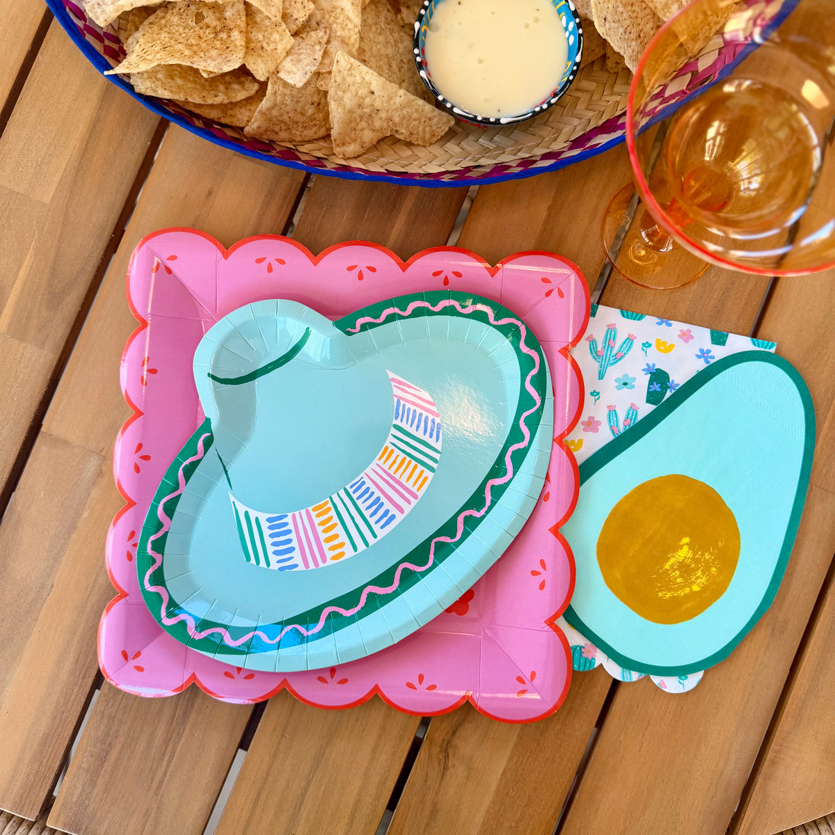 Overhead view of a Cinco de Mayo place setting showing layered pink papel picado and sombrero paper plates with avocado napkin and chips and salsa in the background