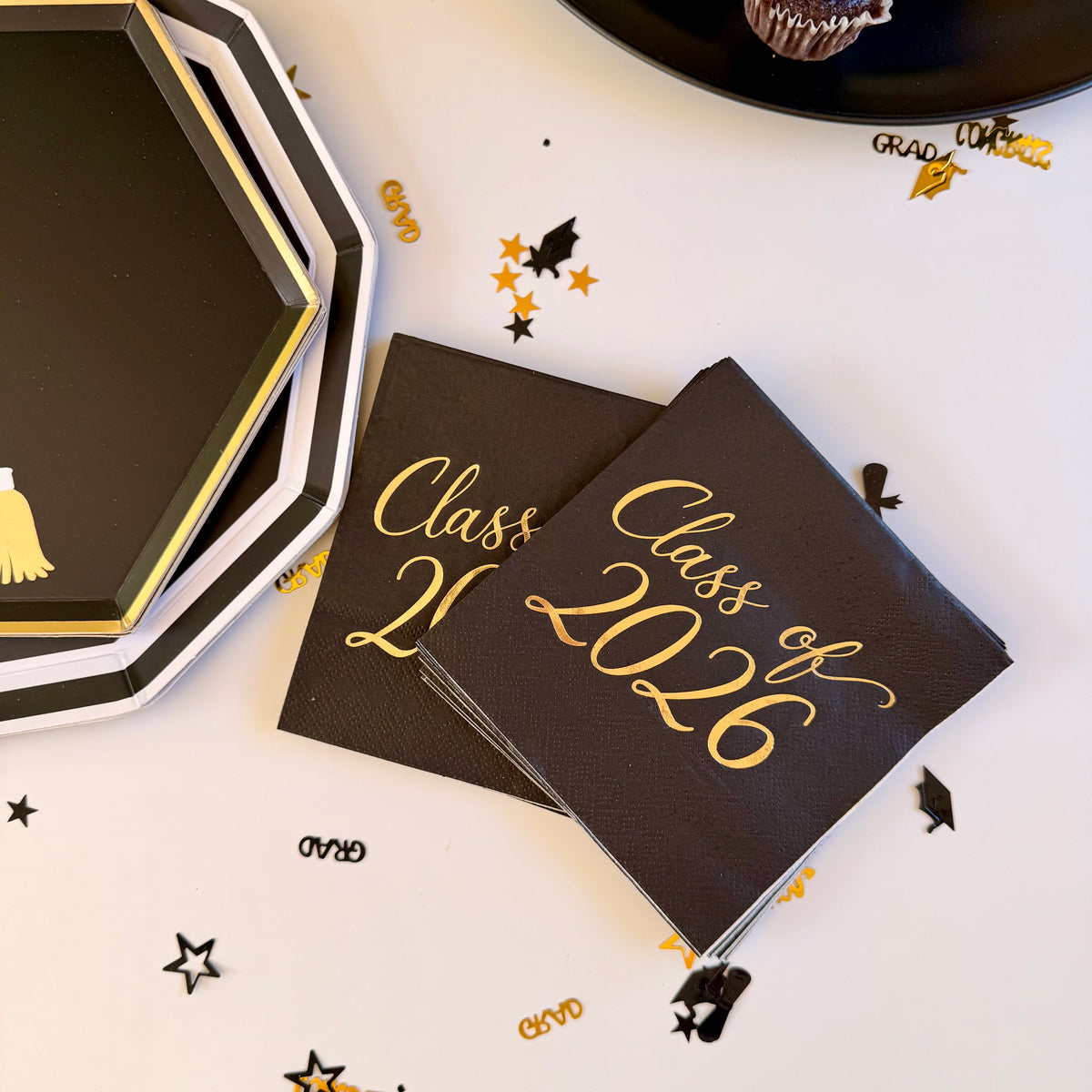 Class of 2026 black cocktail napkins with gold foil script lettering, fanned next to a stack of black octagonal grad cap paper plates and a chocolate cupcake