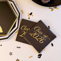 Class of 2026 black cocktail napkins with gold foil script lettering, fanned next to a stack of black octagonal grad cap paper plates and a chocolate cupcake
