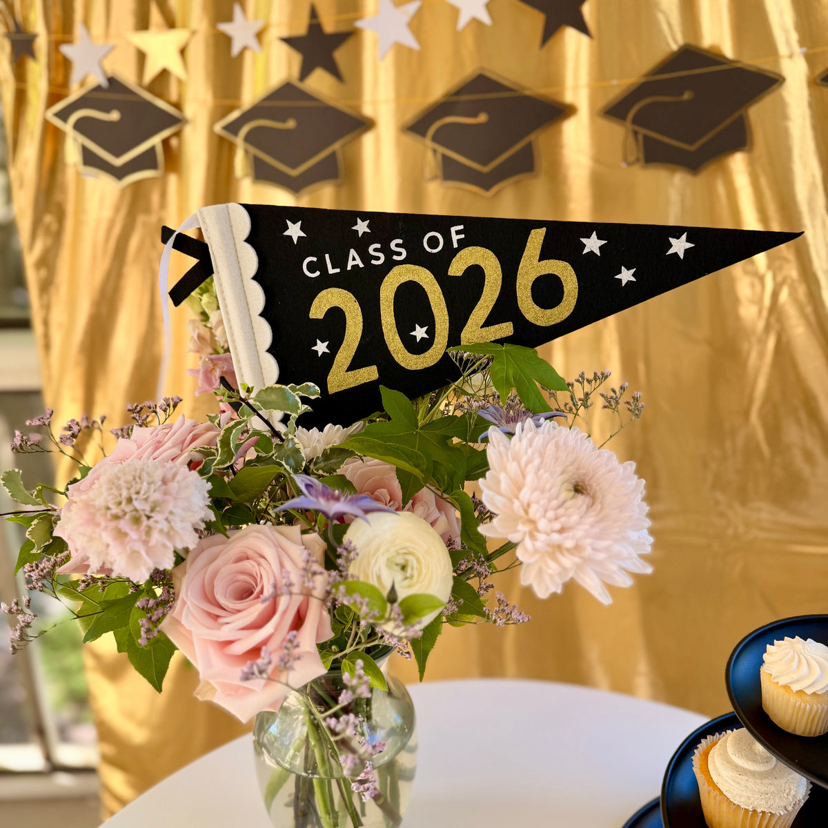 Black and gold Class of 2026 felt pennant flag tucked into a blush and white floral centerpiece, with a grad cap and star banner hanging across a gold backdrop