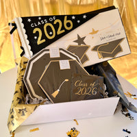 Class of 2026 graduation party kit in a white gift box with black and gold grad cap plates, Class of 2026 cocktail napkins, felt pennant flag, and a star and grad hat banner set