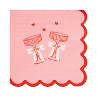 This image shows a pink cocktail napkin with red scalloped edges and an illustration of two clinking coupe glasses tied with bows.
