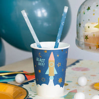 Party cup with rocket design and straw on a festive table with balloons and cake.