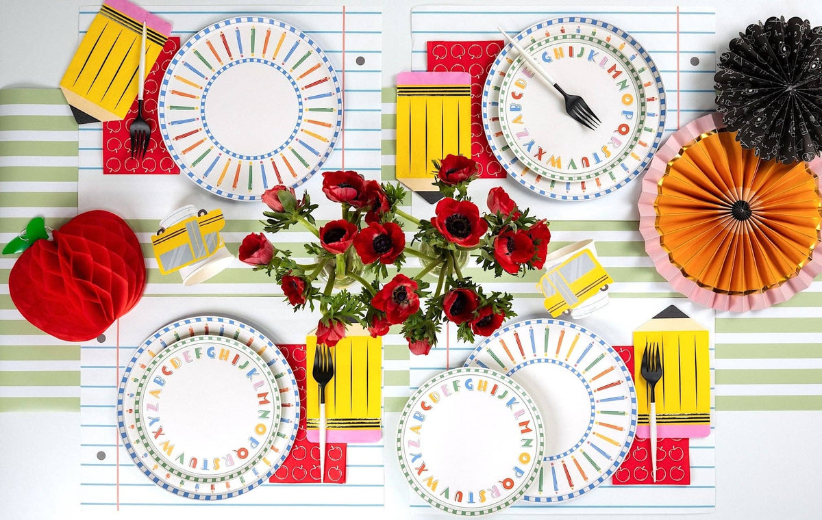 Overhead tablescape with four back-to-school place settings featuring colored pencil paper plates layered under ABC alphabet paper plates, pencil-shaped yellow napkins, math notebook-shaped napkins, red apple-print napkins, school bus napkins, and a central red anemone arrangement