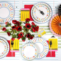 Overhead tablescape with both colored pencil paper plates and ABC alphabet paper plates layered, central red anemone arrangement, school bus paper cup, and pencil-shaped napkins
