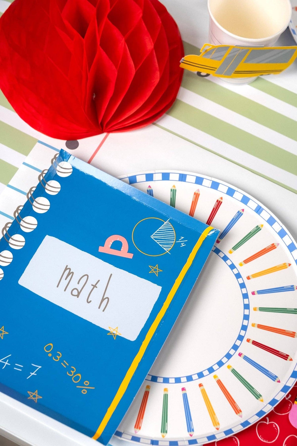Back-to-school place setting featuring a colored pencil paper plate, math notebook-shaped paper napkin, school bus paper cup, red honeycomb apple decoration, on a green-and-white striped paper runner