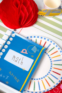 Back-to-school place setting featuring a colored pencil paper plate, math notebook-shaped paper napkin, school bus paper cup, red honeycomb apple decoration, on a green-and-white striped paper runner