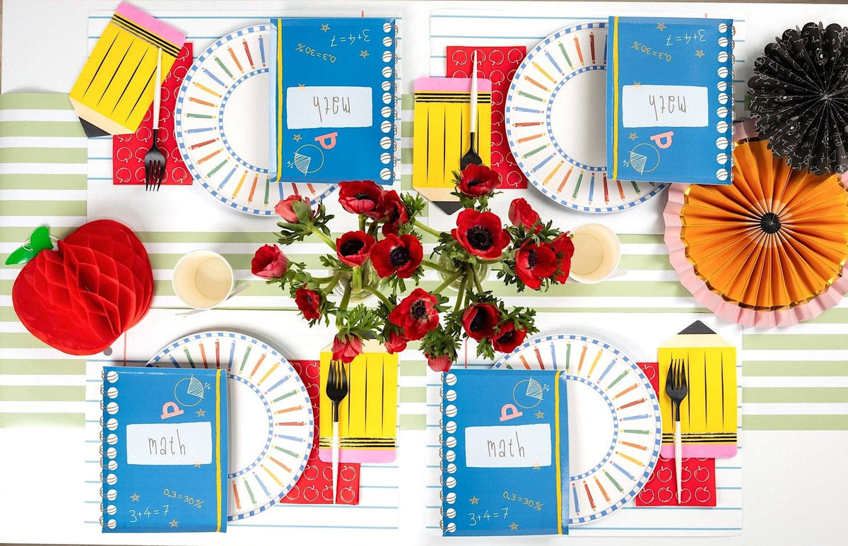 Close-up of a colored pencil paper plate showing the double blue-and-white checkered ring borders and the rainbow-colored pencils arching around the rim, with a math notebook napkin overlapping the edge