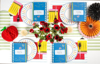 Close-up of a colored pencil paper plate showing the double blue-and-white checkered ring borders and the rainbow-colored pencils arching around the rim, with a math notebook napkin overlapping the edge