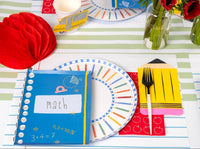 Close-up of a colored pencil paper plate with a math notebook-shaped paper napkin layered on top, school bus paper cup, red apple-print napkin holding black flatware inside a pencil-shaped napkin, on a green-and-white striped runner