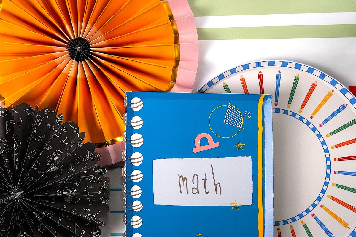 Vertical composition showing math notebook-shaped paper napkin, orange and pink scalloped paper fans with gold accents, black math-equation chalkboard paper fan, and the edge of a colored pencil paper plate