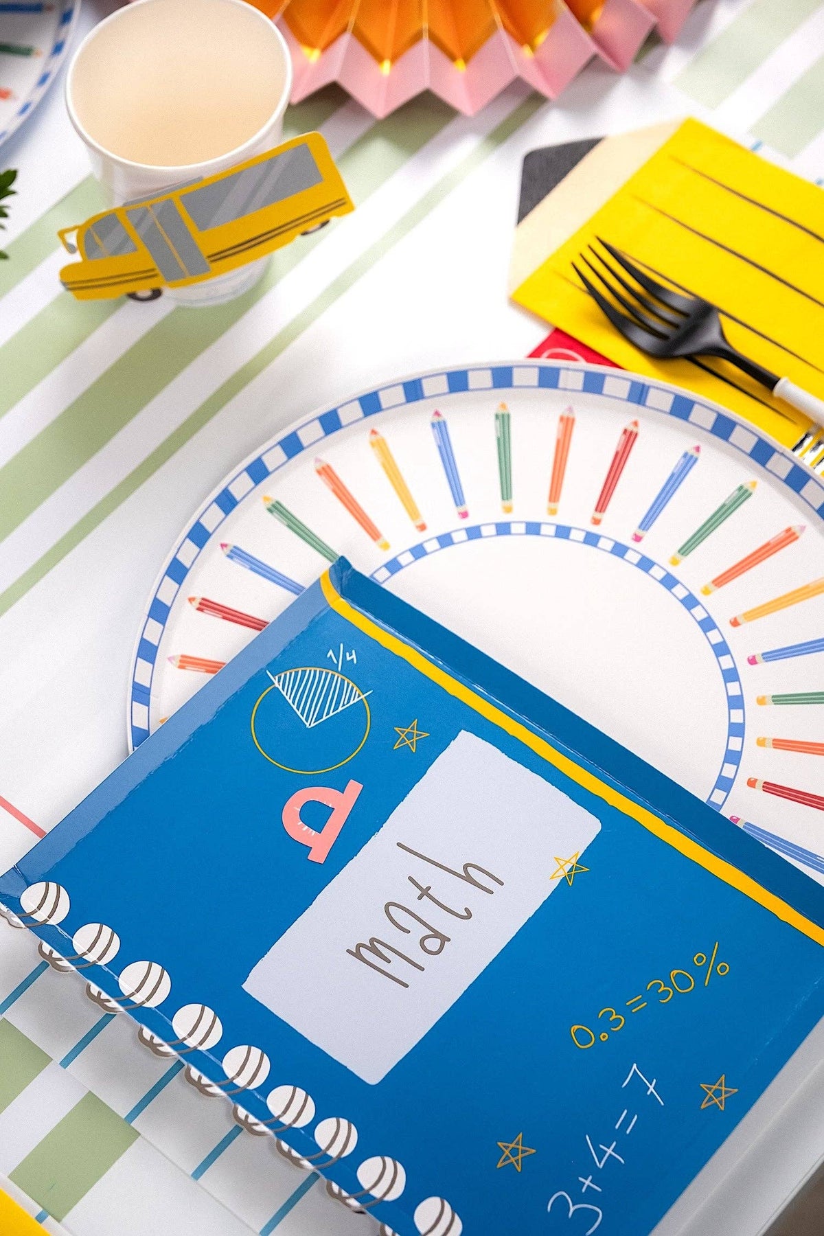 Angled view of a colored pencil paper plate place setting with math notebook-shaped napkin, school bus paper cup, red honeycomb apple, pencil-shaped yellow napkin, on a green-and-white striped runner