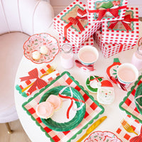 Holiday dessert table with stacked red gingham cookie boxes, cups wrapped with red bows, wreath plates, and Santa cookies on a white round table.