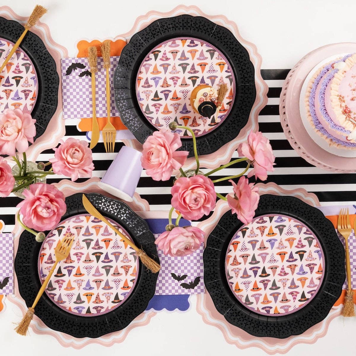 Overhead coquette Halloween table showing multiple place settings with witch hat plates on black chargers, pink roses in vases, and black-and-white striped runner.