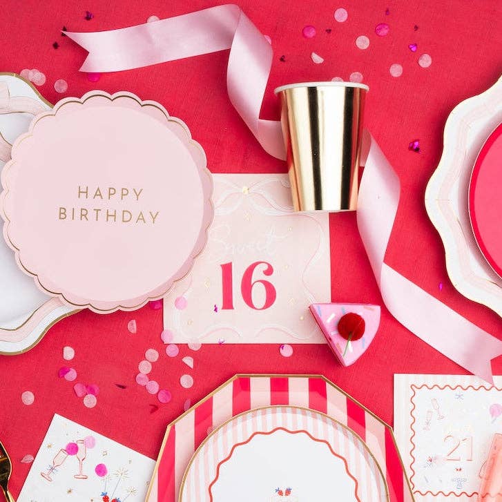 Flat lay of a pink and red birthday table with Sweet 16 bow napkins, scalloped plates, and gold party cups.
