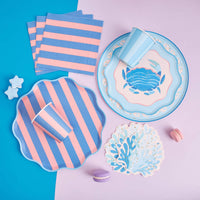 Coastal party flatlay with coral shaped napkins, a blue crab plate, pink and blue striped plates, cups, and napkins.