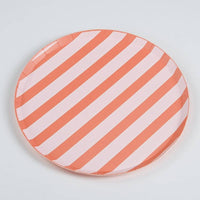 Full coral stripe dinner plate on a white surface, shot from a slightly above angle. Diagonal stripes run across the full face of the plate — approximately 8 alternating coral-orange and pale pink stripes visible. Round plate with clean edge, no scallop. Paper crimping visible at rim.