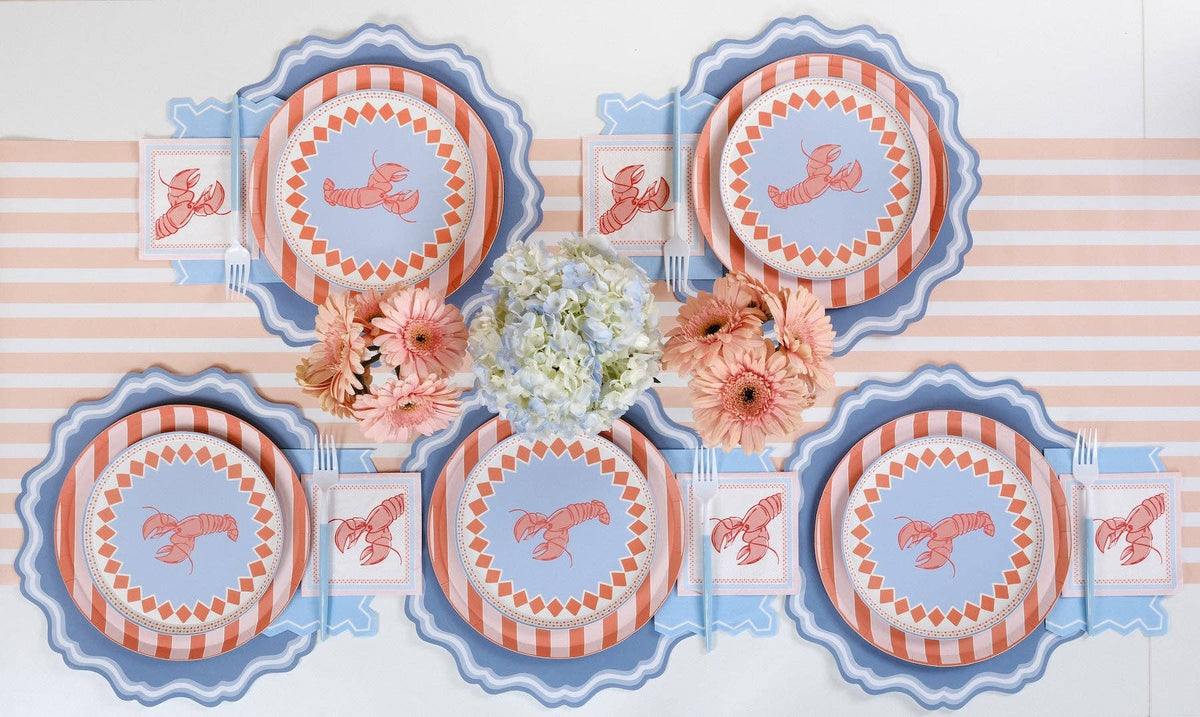 Close-up of one place setting on the blue stripe runner. Coral stripe plate stacked under the lobster salad plate with blue fork and knife resting on the salad plate. Lobster cocktail napkins on both sides. Peach gerberas and white daisies in bud vases.