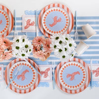 Same angle and setting as Image 6 from a slightly closer vantage. Lobster salad plate and coral stripe plate are clearly layered. Blue fork and knife rest on the salad plate. Lobster napkins visible flanking the place setting. Daisy and gerbera arrangements behind.