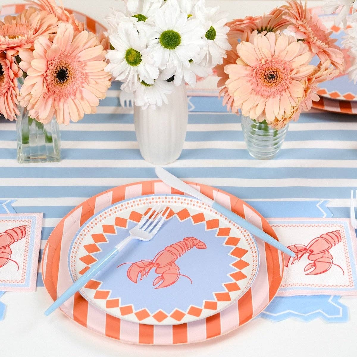 Angled close-up of a place setting on the blue double-wave scalloped placemat over a peach stripe runner. Lobster salad plate on top of the coral stripe dinner plate. The stripe plate's diagonal coral-and-pink pattern is visible around the salad plate edge. Light blue zigzag guest towel under a lobster cocktail napkin to the right. Blue hydrangea and peach gerbera in background.