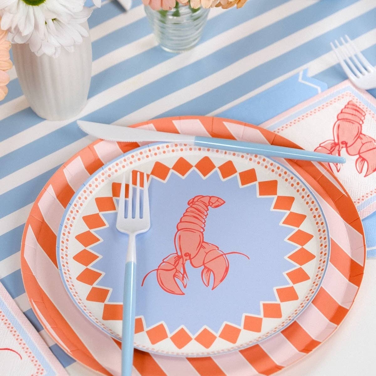 Overhead of a table set for four to six on a blue-and-white stripe table runner. Coral stripe dinner plates under lobster salad plates at every setting. Lobster cocktail napkins flanking each plate. Blue forks, peach gerbera and white daisy centerpieces. White speckled cups at upper right.
