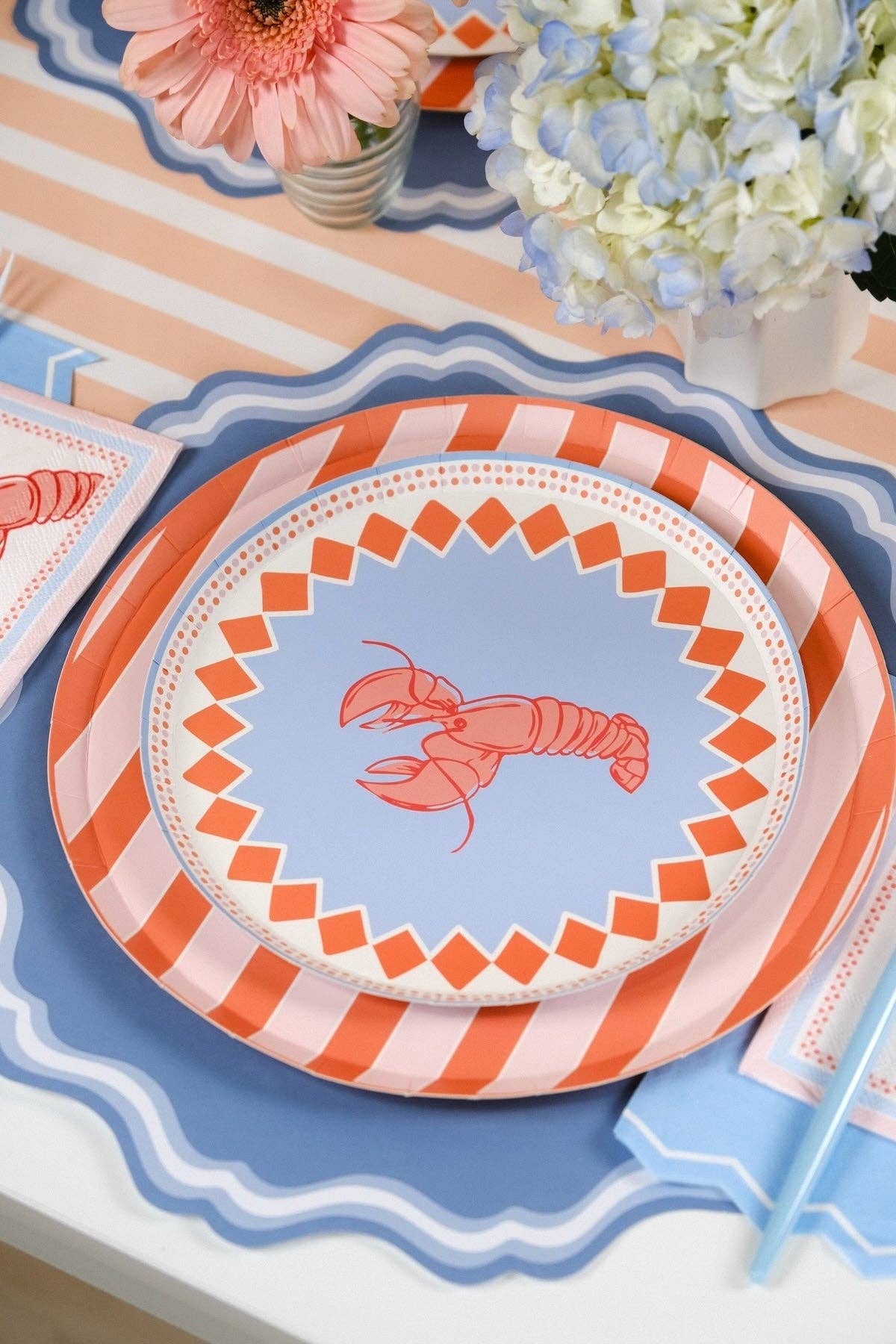 Wide overhead shot of a table set for five with blue double-wave scalloped placemats and a peach-and-white stripe table runner. Each place setting layers the coral stripe dinner plate under the periwinkle blue lobster salad plate. Lobster cocktail napkins and light blue zigzag guest towels at each setting. Peach gerberas and blue hydrangea centerpieces.
