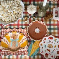 Corn shaped paper napkin with yellow kernels and green husk tucked between a turkey plate and pie plate on a fall plaid tablecloth