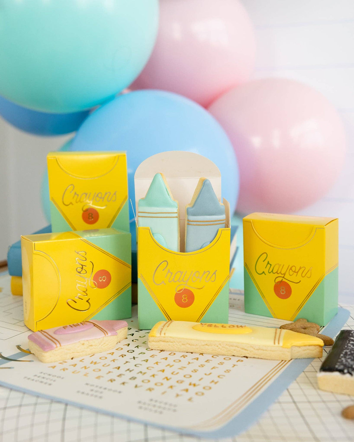 Same balloon backdrop setup as Image 4, slightly wider angle. Five crayon box-shaped treat boxes visible — three upright, one on its side — showing yellow and mint coloring with gold foil script. Crayon-shaped iced sugar cookies in foreground.