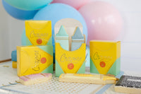 Three assembled crayon box-shaped treat boxes displayed upright against a backdrop of pastel blue and pink balloons. Center box is open showing mint and blue crayon-shaped sugar cookies inside. Two closed boxes flank it. Pink and yellow crayon-shaped cookies are scattered in front on a back-to-school activity sheet.