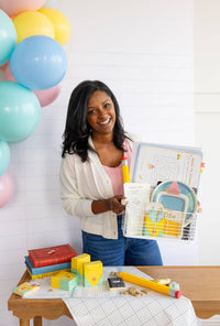 Smiling woman in white cardigan holds a white wire basket filled with back-to-school party supplies including a globe plate, composition notebook plate, library card, and activity book. In the background on a wood table: multiple assembled crayon treat boxes, a novelty pencil, and ABC cookies.