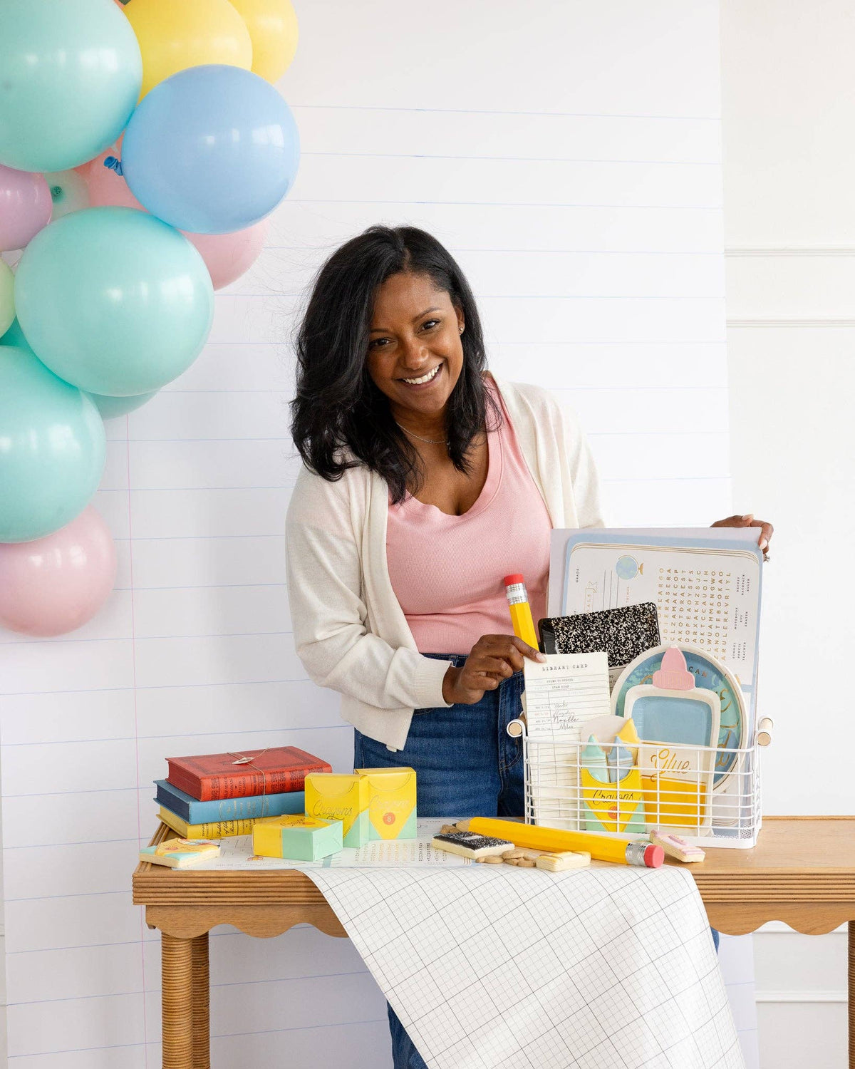 Woman at table holding the back-to-school activity placemat open to show the maze and word search. Assembled crayon treat boxes visible on table left alongside books and novelty pencil.