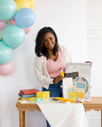 Woman at table holding the back-to-school activity placemat open to show the maze and word search. Assembled crayon treat boxes visible on table left alongside books and novelty pencil.
