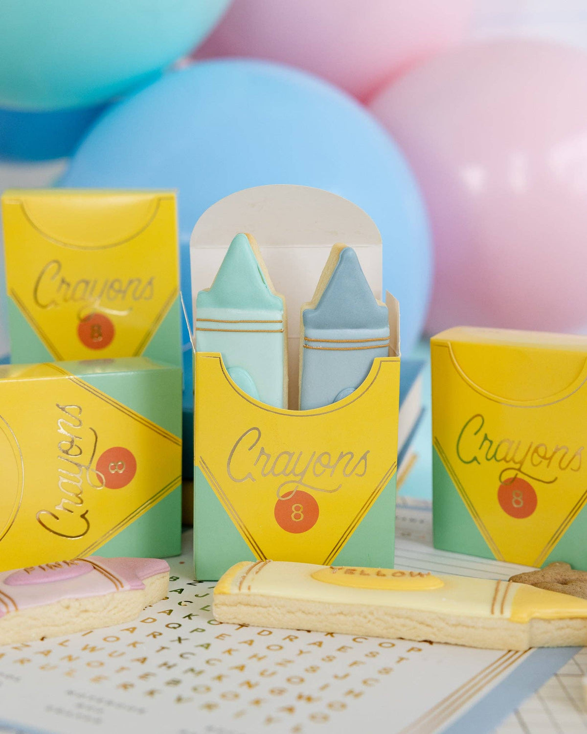Close-up of four assembled crayon treat boxes against pastel balloon backdrop. Center box open showing mint green and blue fondant crayon-shaped sugar cookies inside. Front shows gold foil "Crayons" script detail clearly. Crayon cookies (yellow, pink) in foreground.