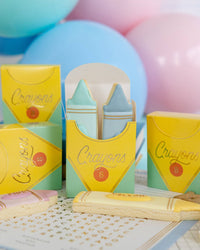 Close-up of four assembled crayon treat boxes against pastel balloon backdrop. Center box open showing mint green and blue fondant crayon-shaped sugar cookies inside. Front shows gold foil "Crayons" script detail clearly. Crayon cookies (yellow, pink) in foreground.