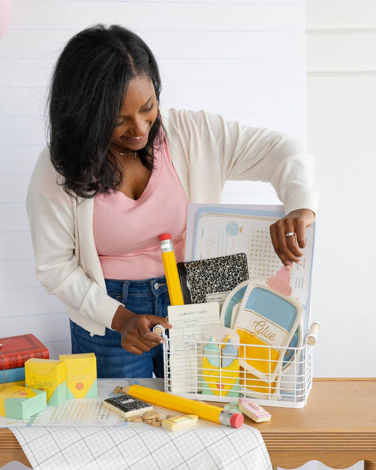 Woman leans over the table arranging the wire basket of back-to-school party goods. Crayon treat boxes and flat-pack boxes visible in foreground lower left. Composition notebook plate, globe plate, and library card visible in basket.