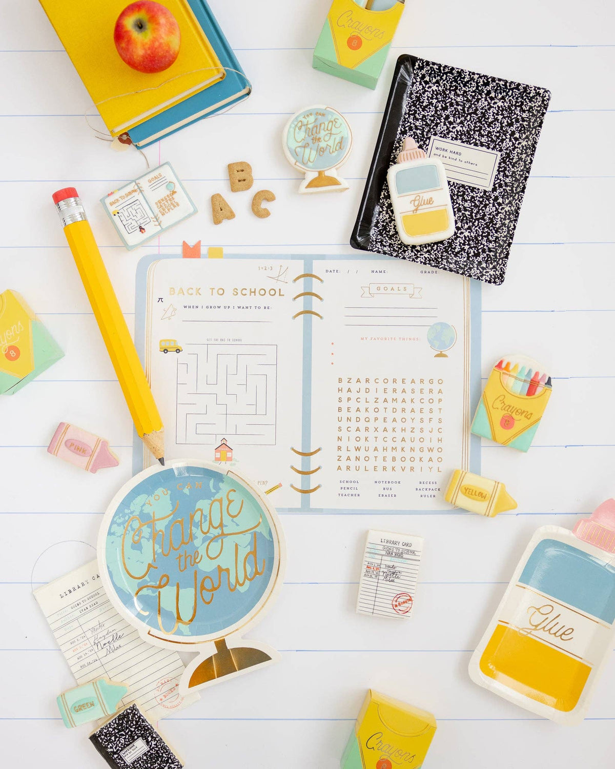 Overhead back-to-school party flat lay on white lined paper background. Assembled yellow-and-mint crayon treat boxes appear in upper right and left corners filled with small treats. Surrounded by a globe plate, composition notebook plate, library card notepads, ABC letter cookies, crayon cookies, a giant novelty pencil, and a back-to-school activity placemat.