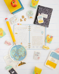 Overhead back-to-school party flat lay on white lined paper background. Assembled yellow-and-mint crayon treat boxes appear in upper right and left corners filled with small treats. Surrounded by a globe plate, composition notebook plate, library card notepads, ABC letter cookies, crayon cookies, a giant novelty pencil, and a back-to-school activity placemat.
