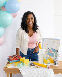 Same woman stands at a wood scalloped-edge table covered with a grid-print white runner. Assembled crayon boxes in yellow and mint visible on the left side of the table alongside vintage books, a novelty pencil prop, mini composition notebooks, and the wire basket of supplies.