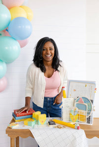 Full-length shot of woman at the same styled table. Crayon treat boxes visible in center-left of frame beside stacked books. White grid-print table runner, pastel balloon arch backdrop.