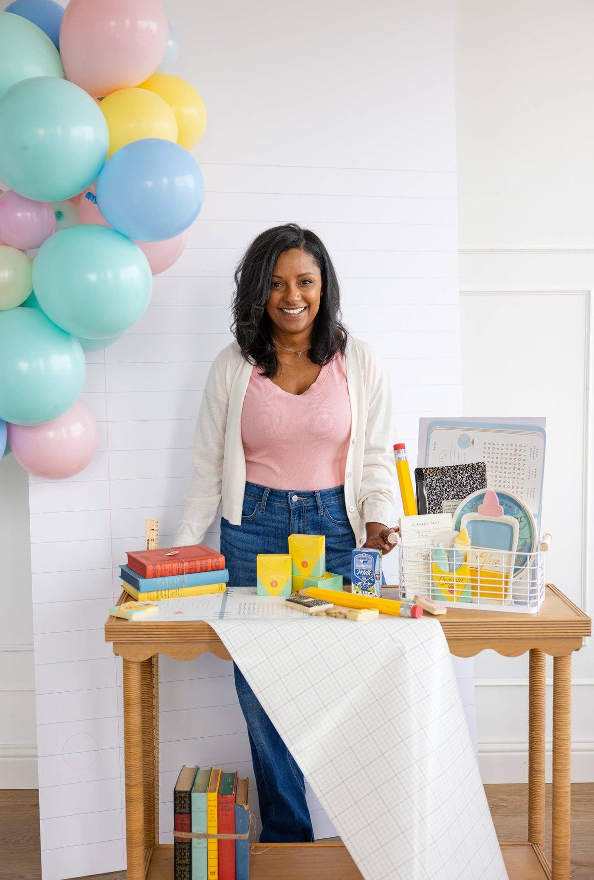 Full table scene with woman standing behind scalloped-edge wood table. Crayon boxes in yellow and mint at center. Books stacked to the left, a small school milk carton visible among props. Grid table runner, white shiplap backdrop.